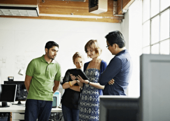 A group of young individuals engaged in a knowledge-sharing session, actively listening to a senior mentor while she uses her iPad, illustrating the process of finding and connecting with influential mentors in your field.