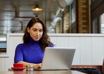 Female SMM intern in a blue top working on a laptop in a cafe, implementing social media marketing strategies.